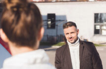 A man with short hair and a beard, wearing a white turtleneck sweater and a dark jacket, stands outdoors with a slight smile. In the foreground, a person with their back to the camera appears to be facing him. The background features a building and some trees.