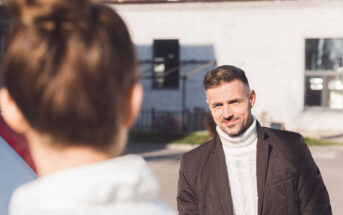 A man with short hair and a beard, wearing a white turtleneck sweater and a dark jacket, stands outdoors with a slight smile. In the foreground, a person with their back to the camera appears to be facing him. The background features a building and some trees.