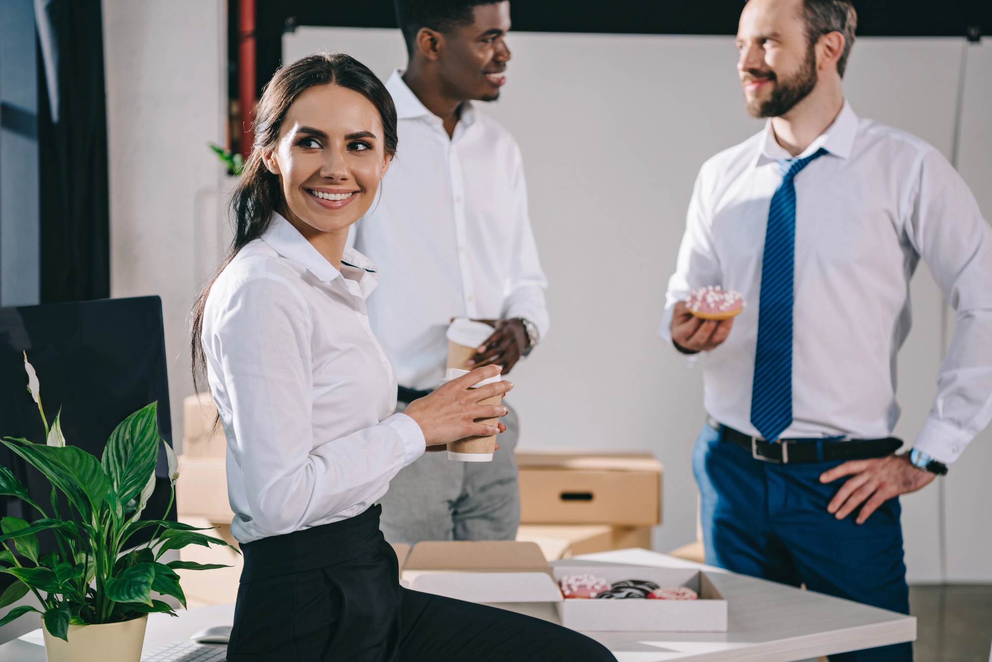 An office scene with three people socializing. A woman in a white blouse and black pants holds a coffee cup, smiling as she sits on a desk. Two men, also in business attire, stand nearby chatting. One holds a coffee cup, and the other holds a donut. A plant is on the desk.