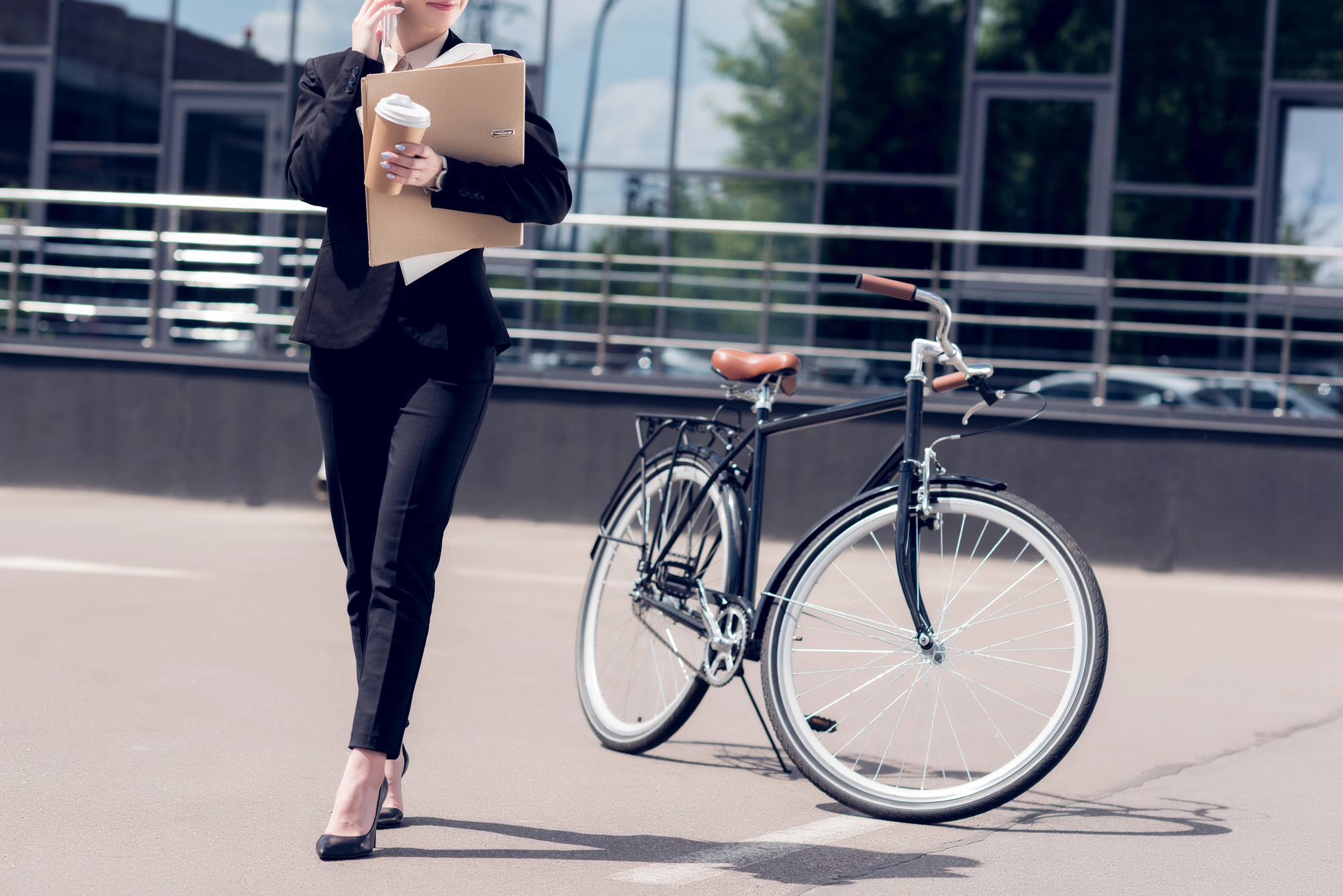 A person in business attire holding a coffee cup and folder is talking on the phone while walking past a bicycle. The person is in front of a modern building with large windows. The bicycle is parked nearby, and the environment appears urban.