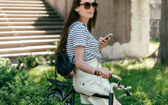 A woman with long hair, wearing sunglasses, a striped shirt, and a light-colored skirt is sitting on a bicycle while holding a smartphone. She is outdoors near some steps and greenery, looking to the side with a small backpack on her back.