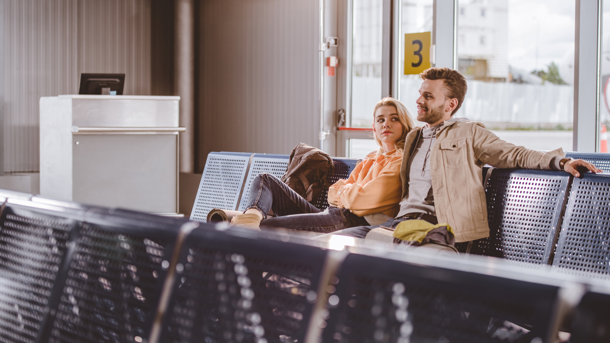 Two people sitting together on blue chairs in an airport waiting area. They appear relaxed, with one person resting their arm on the back of the seat. In the background, there is a gate marked with a "3" and a counter. Sunlight filters through the large windows.