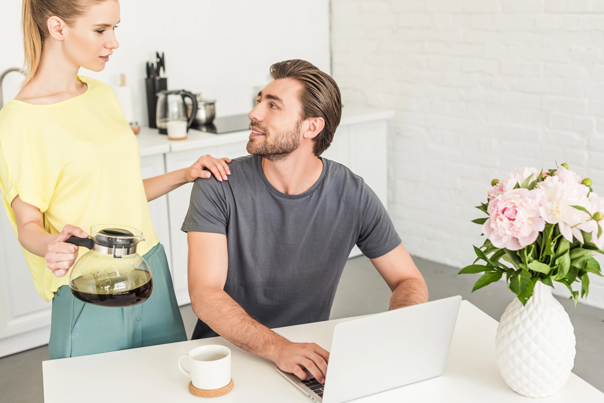 A man is sitting at a table with a laptop in front of him, looking up at a woman who is standing beside him pouring coffee. The woman has one hand on the man's shoulder. A white vase with pink flowers is on the table. They are in a bright kitchen.