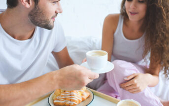 A man and woman in casual clothing sit closely together, enjoying a breakfast in bed. The man holds a cup of coffee on a saucer towards the woman, who is smiling. A tray with a croissant is placed on the bed in front of them.