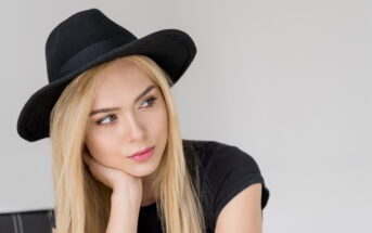 A young woman with long blonde hair wears a black hat and a black t-shirt. She rests her chin on her hand and gazes thoughtfully to the side. The background is light gray and unobtrusive.