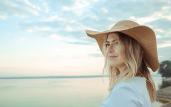 A woman with shoulder-length blonde hair is wearing a wide-brimmed hat and looking off into the distance. She stands by a calm body of water under a cloudy sky during what appears to be either sunrise or sunset. The background features a peaceful and serene landscape.