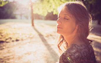 A woman with long hair stands outdoors with her eyes closed while the sun shines through the trees in the background. She appears to be enjoying the warmth and tranquility of the moment. The light creates a soft glow around her face.