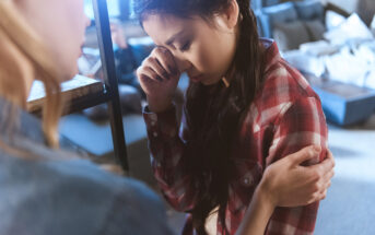 In a softly lit room, a person with long dark hair is holding their head in distress while another person, facing away from the camera, gently holds their arm in a comforting manner. The background shows a blurred living space with furniture.