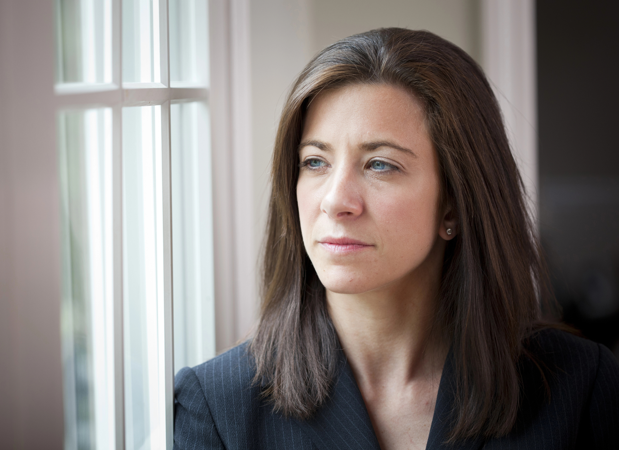 A woman with long brown hair and wearing a dark blazer gazes thoughtfully out of a window. Natural light illuminates her face, highlighting her contemplative expression.