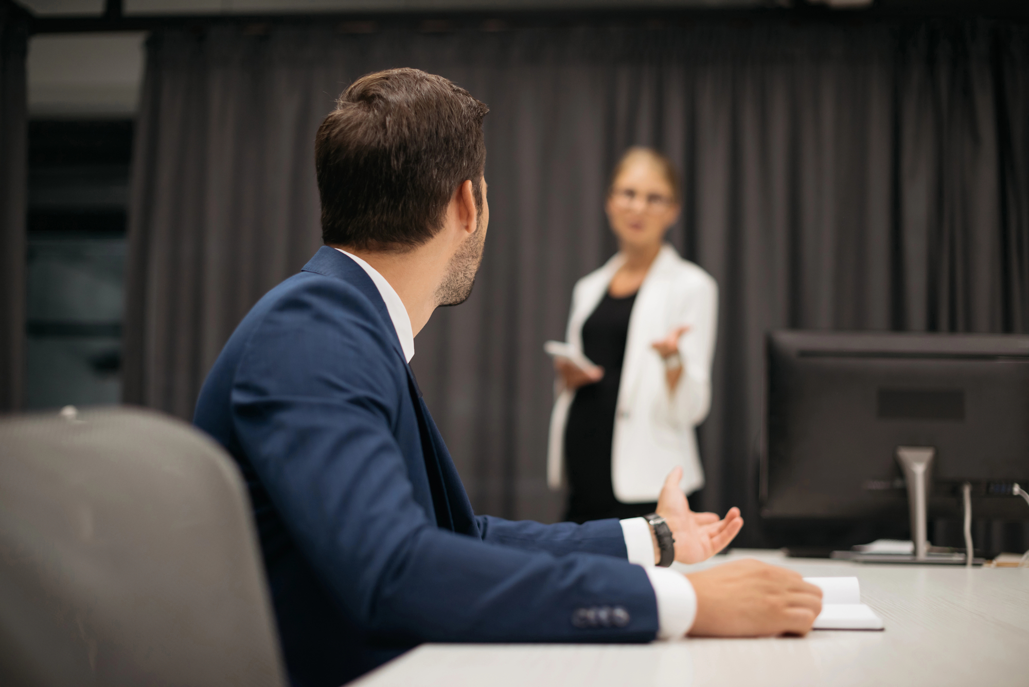 A man in a blue suit is sitting at a desk in an office, turned towards a woman in a white blazer who is standing and talking. A computer monitor is on the desk, and a gray curtain is in the background. The man appears to be gesturing with his hands.