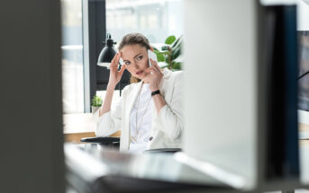 A woman dressed in white sits at a desk in an office, holding her temples and looking stressed. She has long, brown hair tied back, and the desk features a lamp and plant. The background includes large windows and a computer monitor.