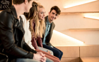 A group of four young adults sit on a stylish, illuminated staircase, engaged in lively conversation. They appear casual and happy, with warm lighting enhancing the cozy atmosphere. The person on the far right is looking directly at the camera, smiling.