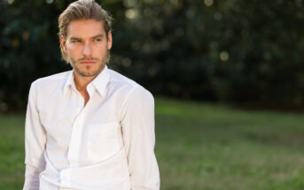 A man with light brown hair, styled back, has a thoughtful expression on his face. He is wearing a white button-up shirt and is sitting outdoors on a green lawn with a blurred, dark green, leafy background.