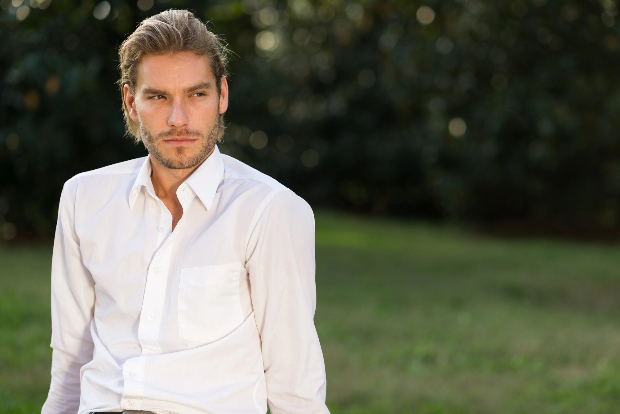 A man with light brown hair, styled back, has a thoughtful expression on his face. He is wearing a white button-up shirt and is sitting outdoors on a green lawn with a blurred, dark green, leafy background.