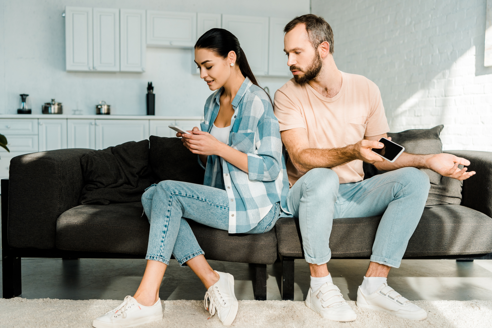 A woman in a blue checkered shirt and jeans is using her smartphone on a couch. A man in a peach shirt and jeans, sitting next to her, glances at her phone while holding his own phone. They are in a modern, bright living room with a kitchen in the background.