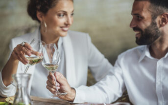 A woman and a man, both dressed in white outfits, are seated at a table and clinking wine glasses together, smiling warmly at each other. The table has some food and a decanter of wine. The background is softly blurred.