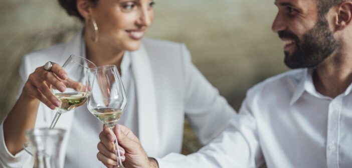 A woman and a man, both dressed in white outfits, are seated at a table and clinking wine glasses together, smiling warmly at each other. The table has some food and a decanter of wine. The background is softly blurred.