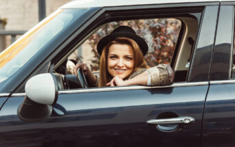 A smiling woman with long hair is sitting in the driver’s seat of a black car with the window down. She is wearing a black hat and a beige plaid shirt, resting her right elbow on the car window frame. The background is blurred with trees and a building.