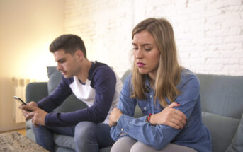 A man and a woman sit on a couch indoors, both appearing upset. The man, looking at his phone, has a neutral expression, while the woman looks down with her arms crossed, seeming distressed. The background includes a white brick wall and a coffee table in front of them.