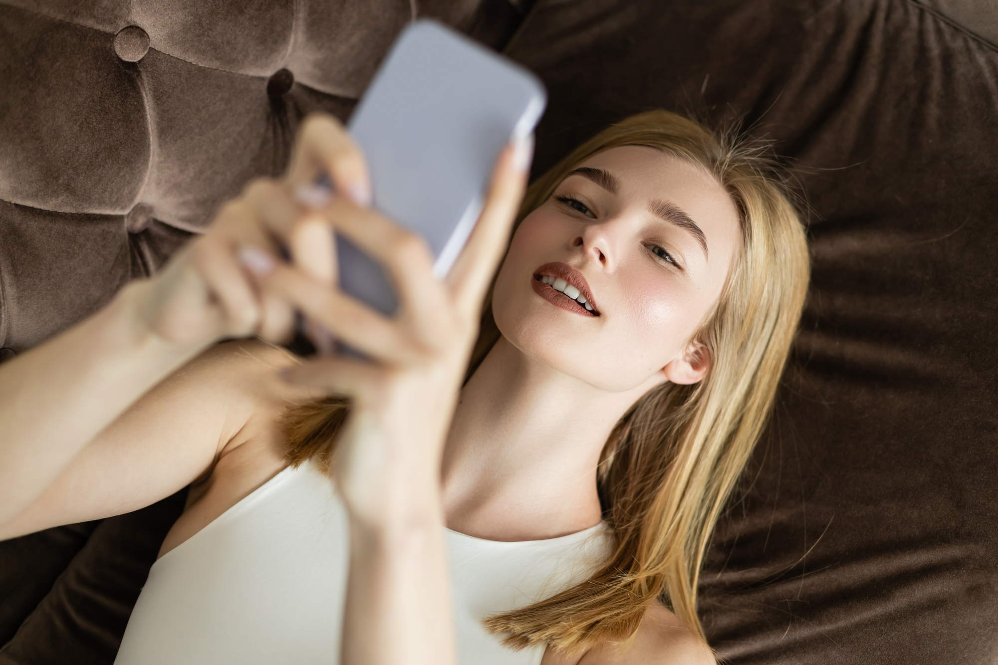 A woman with light brown hair is lying on a dark brown, tufted sofa, holding a smartphone. She is wearing a white sleeveless top and smiling as she looks at the phone screen. The image focuses on her face and the phone.