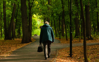 An elderly person walks on a paved path through a serene, green forest. They are wearing a blue jacket and carrying a black bag, with fallen leaves scattered on the ground. Tall trees border the path, creating a peaceful, natural setting.