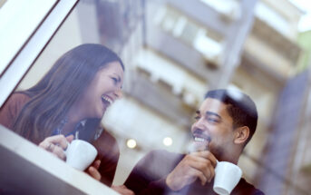 Two people sit at a café table, each holding a coffee cup and smiling warmly at each other. The image is viewed through a window, with soft reflections of lights and surrounding buildings. Both individuals appear to be enjoying their conversation.