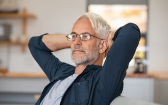 A mature man with gray hair and beard, wearing glasses and a dark shirt, is seated indoors. He has his hands behind his head, looking relaxed and pensive. The background shows blurred kitchen shelves and a window.