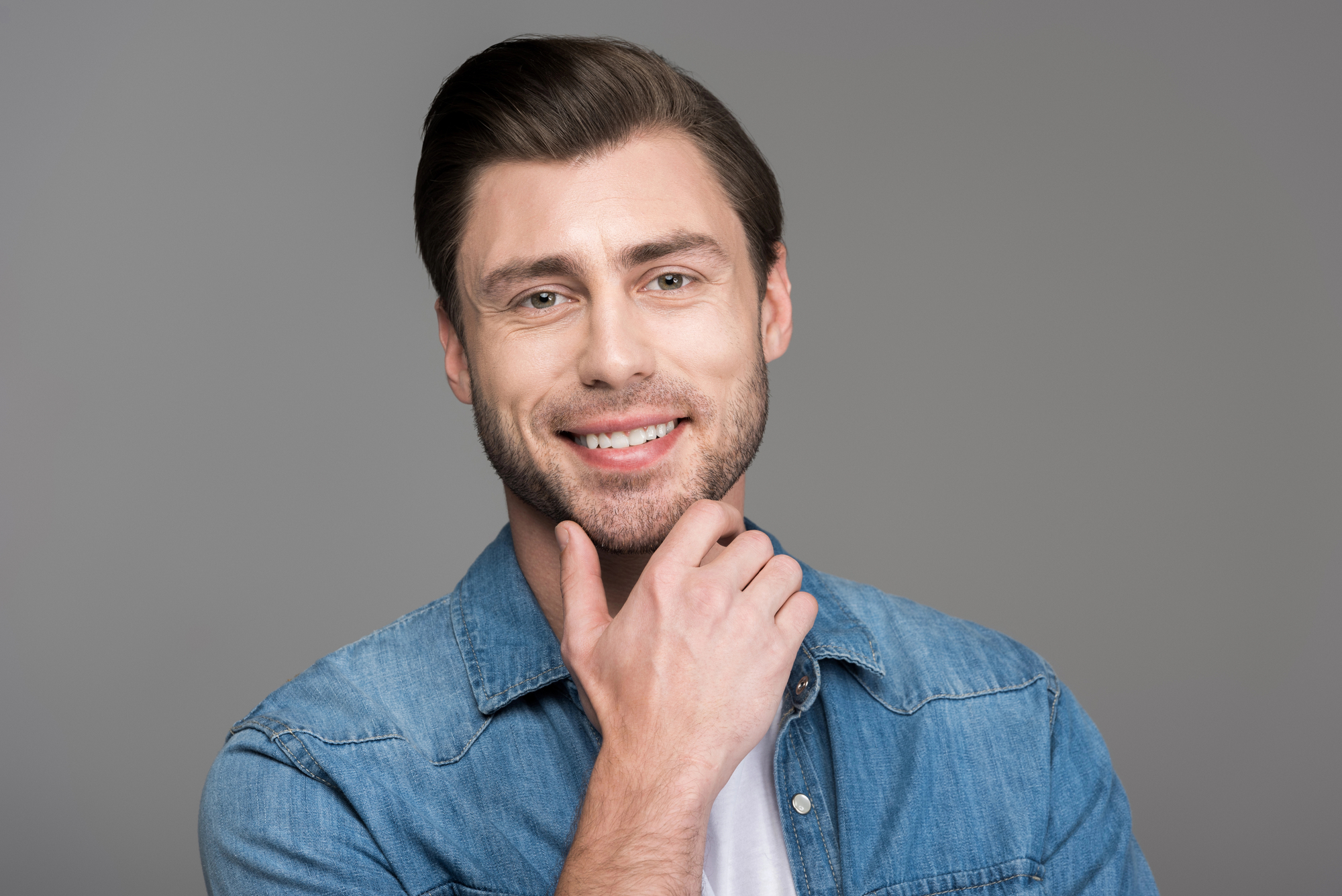 A smiling man with short brown hair and a beard gently touches his chin with his right hand. He is wearing a blue denim shirt over a white T-shirt and stands against a solid gray background.