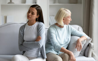 A young woman and an older woman sit back-to-back on a couch, both looking away from each other with serious expressions. The younger woman has dark hair and is crossing her arms, while the older woman, with light hair, rests her hand on the back of the couch.