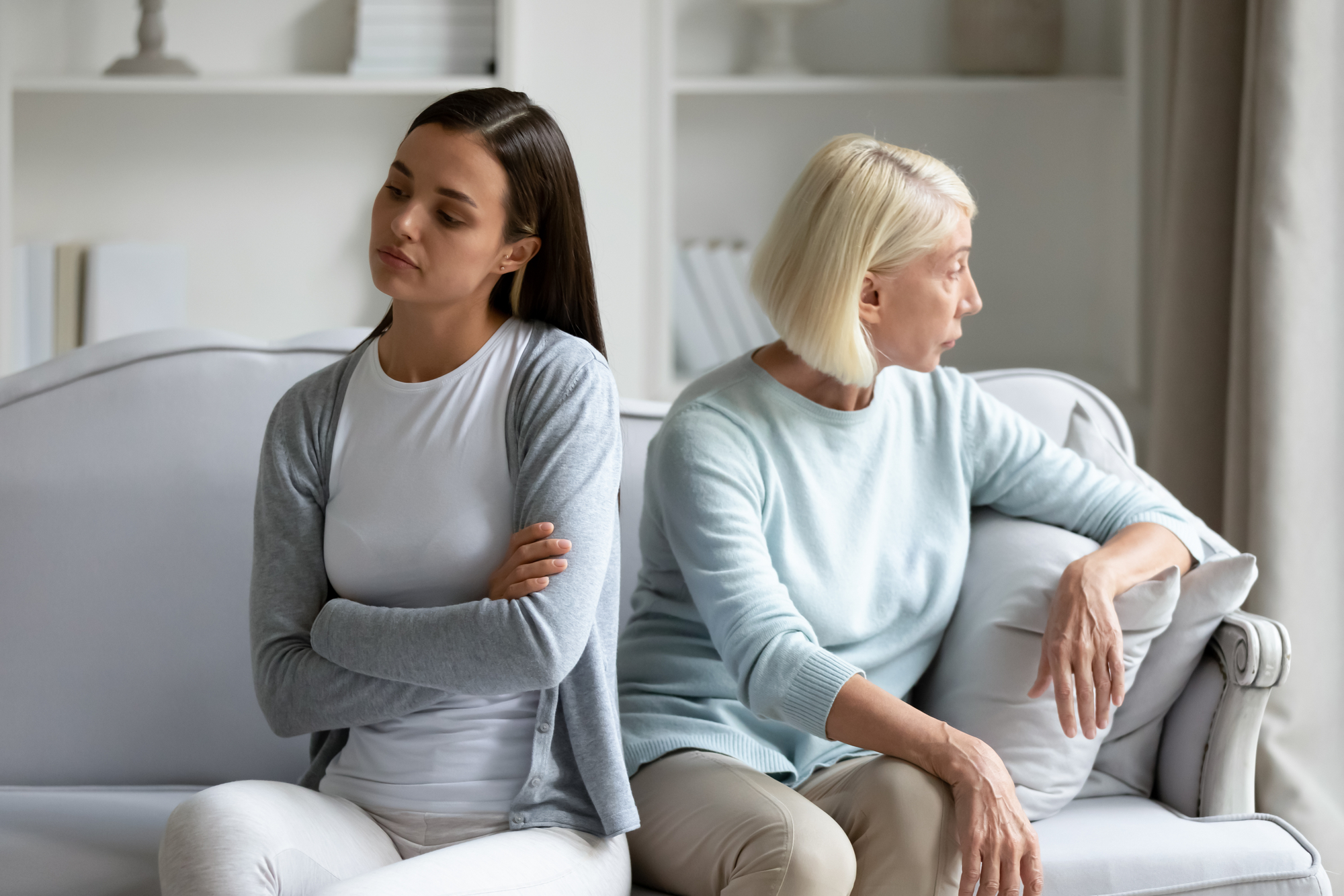 A young woman and an older woman sit back-to-back on a couch, both looking away from each other with serious expressions. The younger woman has dark hair and is crossing her arms, while the older woman, with light hair, rests her hand on the back of the couch.