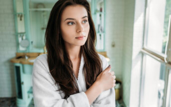 A woman with long brown hair stands in a brightly lit bathroom near a window, wearing a white bathrobe. She looks thoughtfully out the window with one arm crossed over her chest and the other resting on her shoulder. The background includes a vanity and a mirror.