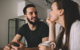 A man with a beard and a woman with long hair sit at a table, smiling at each other. The man is holding a smartphone while the woman is holding a white coffee mug. They are in a well-lit, modern room with minimalistic decor.