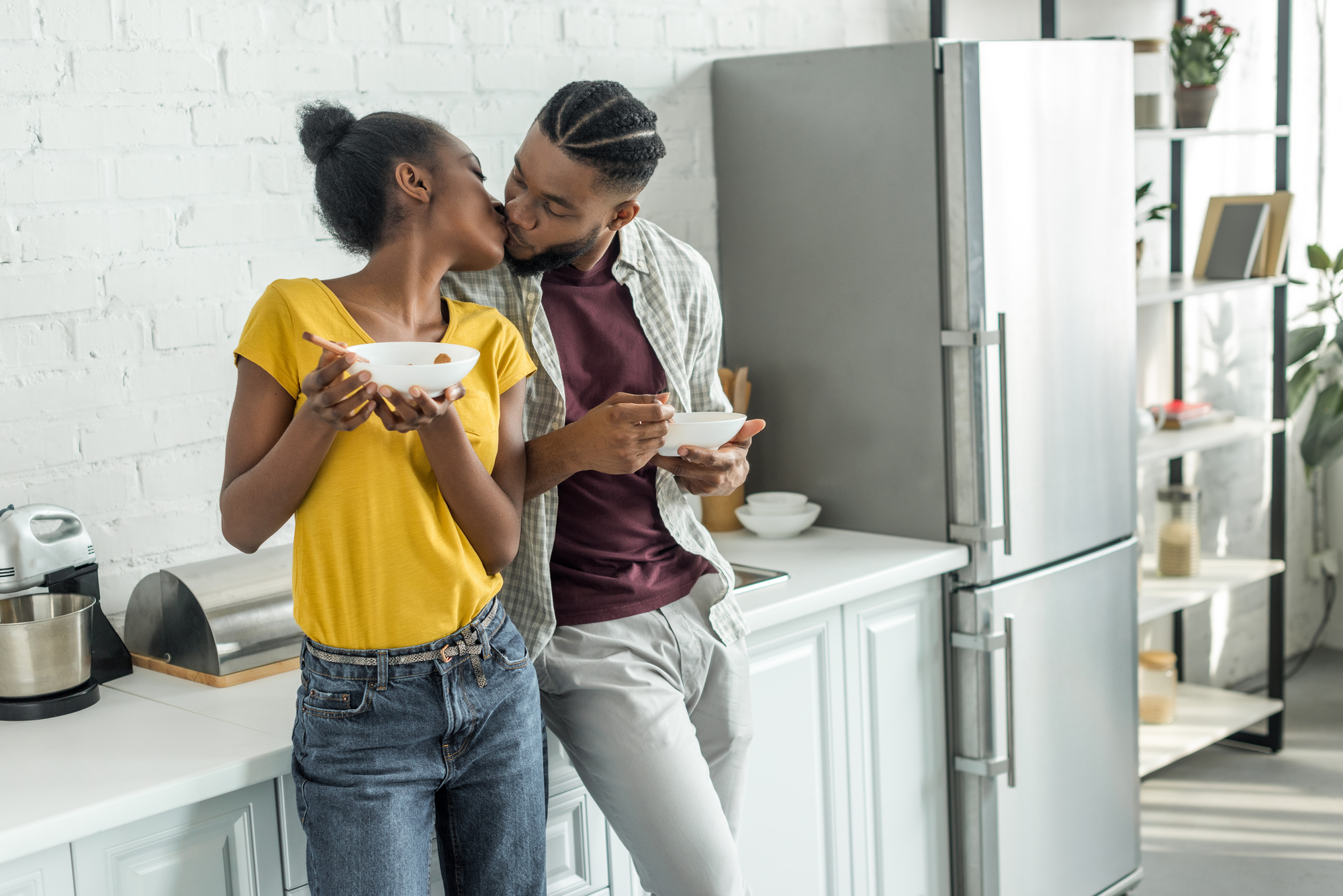 A couple stands in a modern kitchen, sharing a kiss while holding bowls of food. The woman, in a yellow shirt and jeans, stands next to a man in a plaid shirt and jeans who is leaning against the counter. A refrigerator and kitchen items are visible in the background.