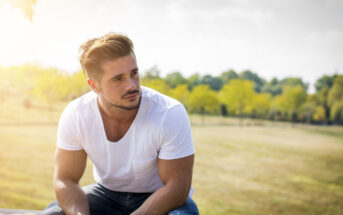 A man with short hair sits on a bench outdoors in a sunny park. He is wearing a white T-shirt and blue jeans, looking pensively into the distance. Green trees and soft sunlight create a peaceful background.