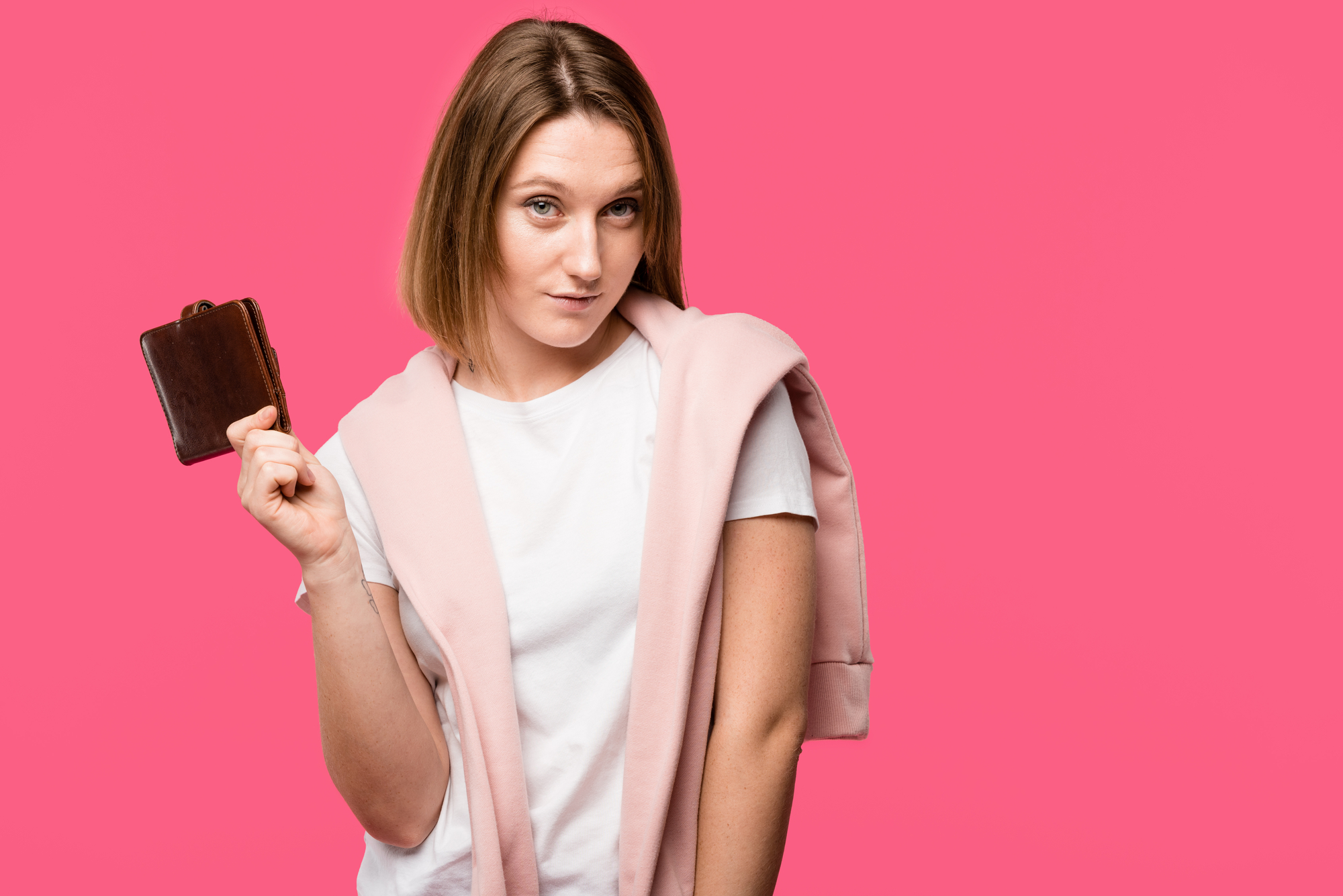 A woman with a light pink jacket draped over her shoulders and wearing a white t-shirt holds up a closed brown wallet in her left hand. She has a neutral expression, and the background is a solid bright pink.