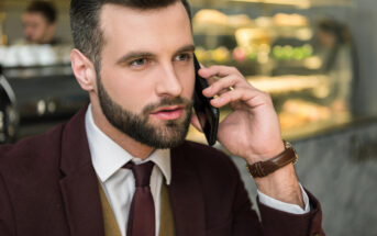 A man with a beard, dressed in a maroon suit and tie, is holding a smartphone to his ear. He is seated, likely in a café or restaurant, with blurred background elements suggesting pastry displays and another person in the distance.