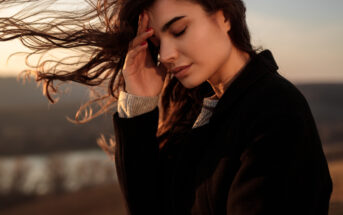 A woman with long, flowing hair stands outdoors with her eyes closed, gently touching her forehead. She is dressed in a black coat. The blurry background shows a serene landscape at sunset. Her expression is peaceful and contemplative.