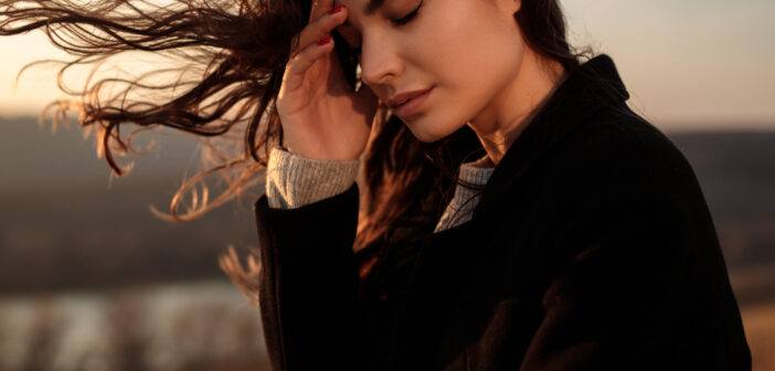 A woman with long, flowing hair stands outdoors with her eyes closed, gently touching her forehead. She is dressed in a black coat. The blurry background shows a serene landscape at sunset. Her expression is peaceful and contemplative.