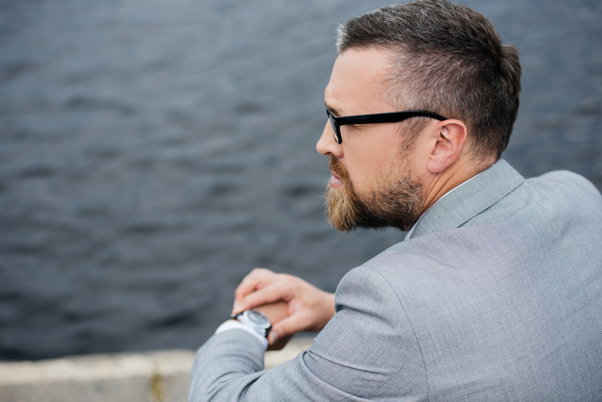 A man with a beard and glasses, dressed in a light gray suit, is leaning on a stone railing while gazing contemplatively over the water. The water is dark and slightly rippled, indicating it's an overcast day. The man is seen from behind and the side.