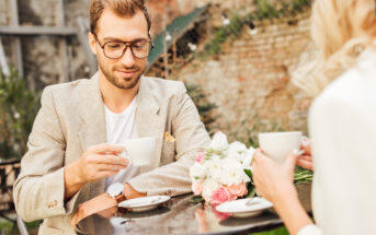 A man with glasses and a beard wearing a beige blazer is sitting at a cafe table outdoors, holding a cup of coffee. Across from him is a woman with blonde hair holding a cup. The table is adorned with a bouquet of flowers. A stone wall and string lights are in the background.