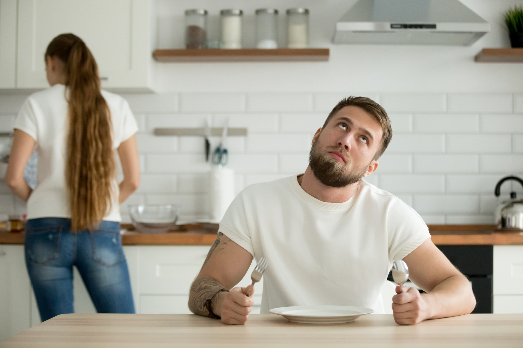 A man with a beard sits at a kitchen table holding a fork and spoon, looking up in anticipation, with an empty plate in front of him. In the background, a woman with long hair, wearing a white shirt and jeans, is busy at the kitchen counter.