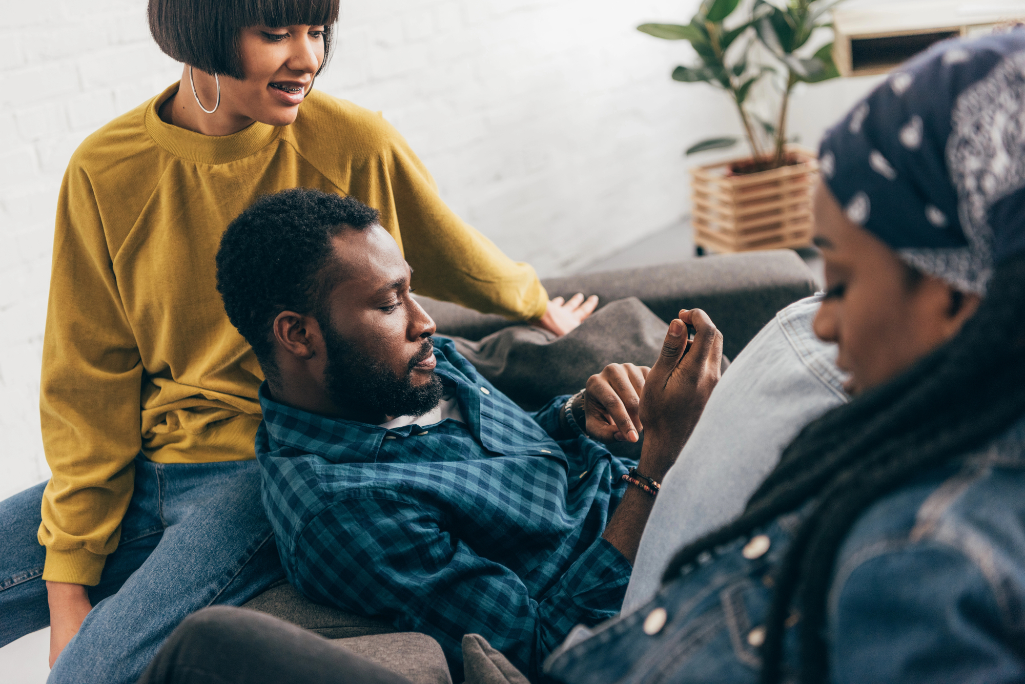 Three friends are relaxing on a couch. The person in the middle is lying back, holding a phone and looking at it, while the person on the left, wearing a yellow sweater, is sitting on the armrest, leaning towards them. The third person on the right is wearing a bandana.