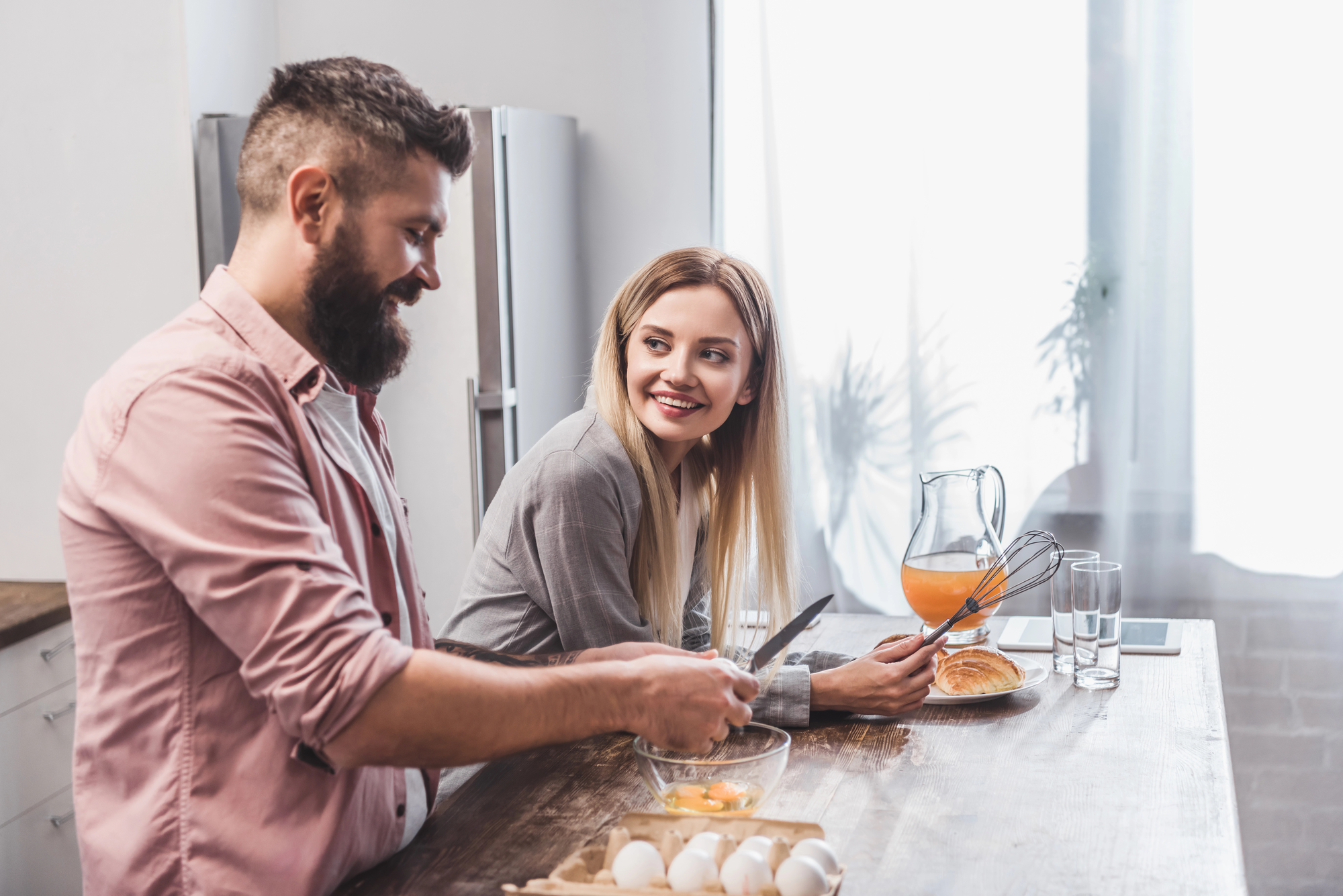 A man and a woman are standing in a kitchen by a wooden table. The man, with a beard and wearing a pink shirt, cracks eggs into a bowl. The woman, with long blonde hair and a grey shirt, smiles at him. On the table are eggs, a croissant, orange juice, and glasses.