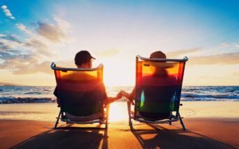 A couple sits on colorful beach chairs, holding hands and facing the sunset. They are on a sandy beach with the ocean waves gently lapping in the background. The sky is a mix of warm orange and yellow hues, reflecting on the water and creating a serene atmosphere.