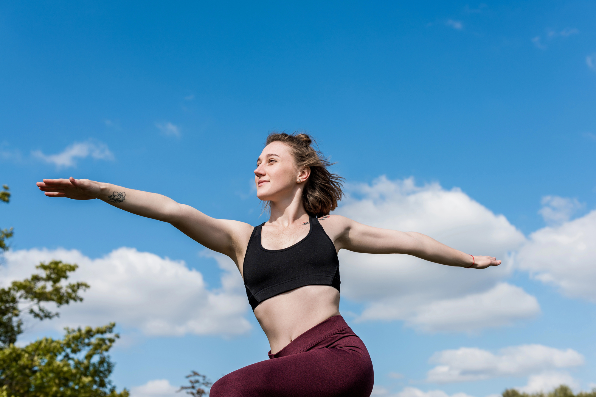 A woman practices a yoga pose outdoors on a sunny day with a clear blue sky and clouds in the background. She is wearing a black sports bra and maroon leggings, balancing on one leg with her arms extended, looking peaceful and focused.