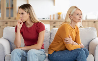 Two women sit on a couch with their bodies turned away from each other, appearing upset. The woman on the left, with long brown hair, wears a red shirt and looks pensive. The woman on the right, with blonde hair, wears a yellow shirt with her arms crossed.