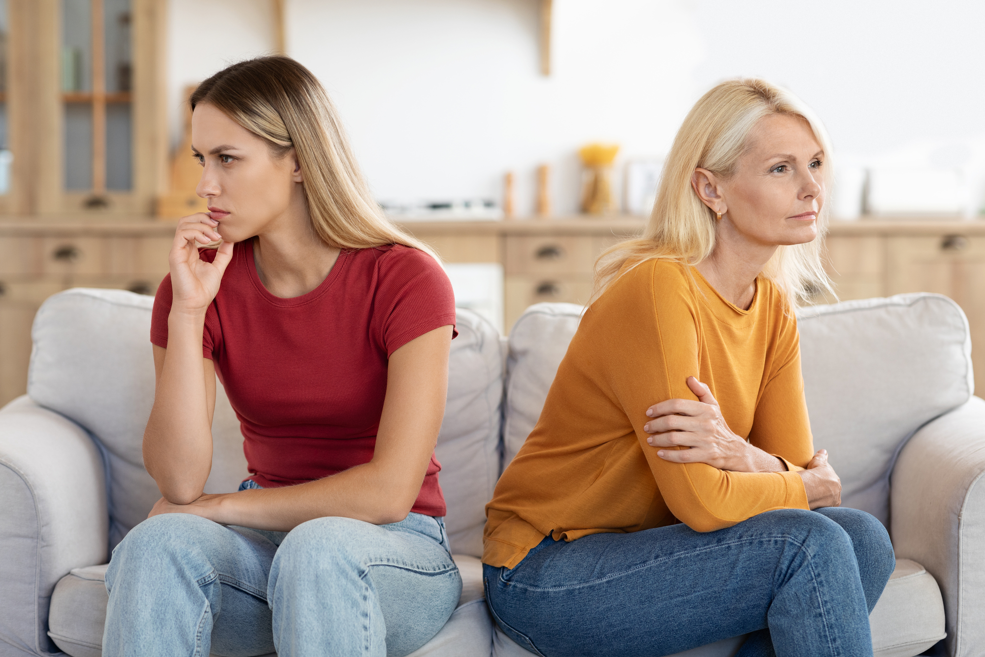 Two women sit on a couch with their bodies turned away from each other, appearing upset. The woman on the left, with long brown hair, wears a red shirt and looks pensive. The woman on the right, with blonde hair, wears a yellow shirt with her arms crossed.