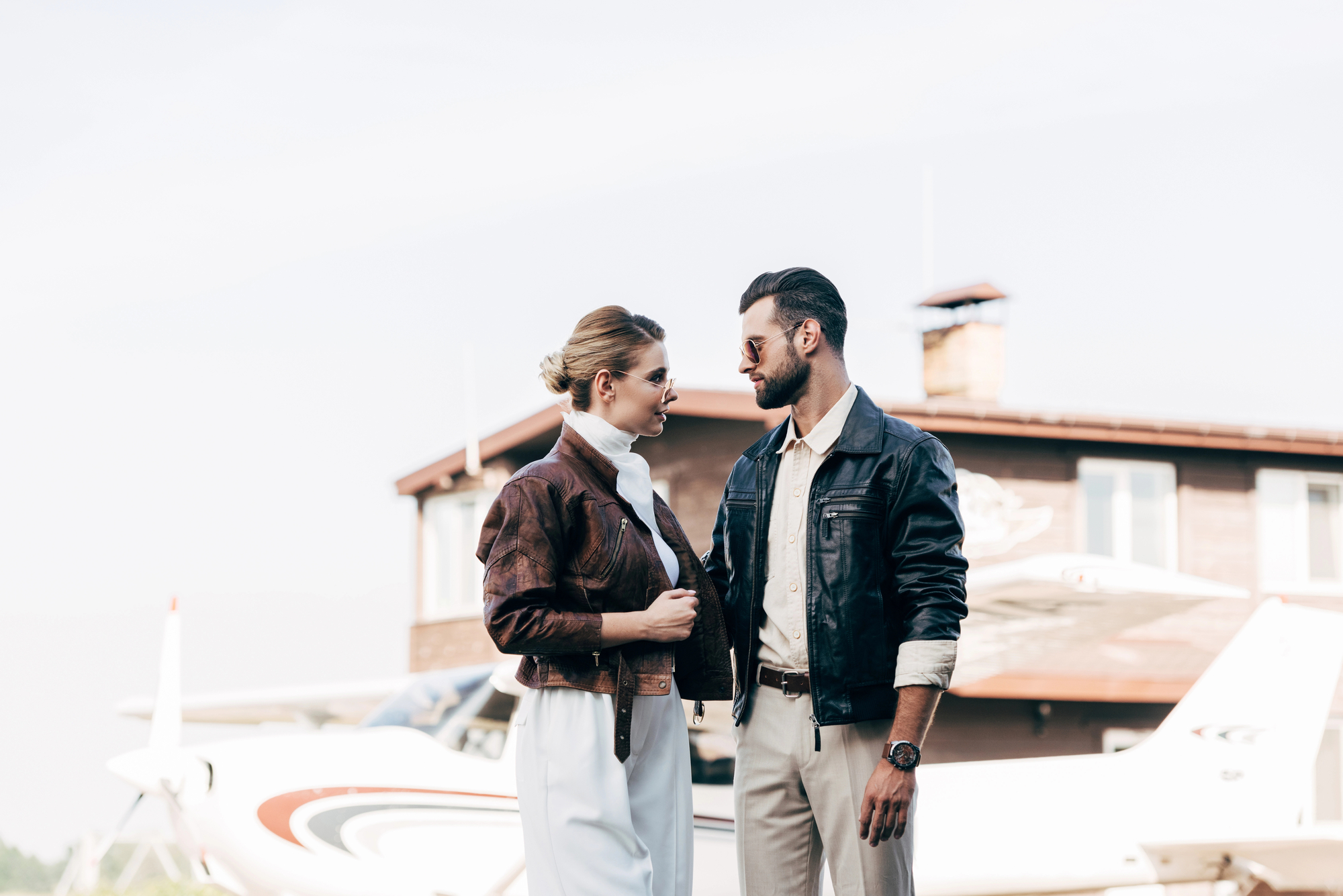 A man and woman, both wearing aviator jackets, stand facing each other near a small airplane with a building in the background. The woman has her hair tied back and is wearing a white outfit. The man has a beard and is dressed in light pants with a dark jacket.