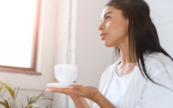 A woman with long dark hair wearing a white shirt is holding a white cup and saucer. She is gazing out of the window, bathed in soft natural light. In the background, there is a green plant and a white wall.