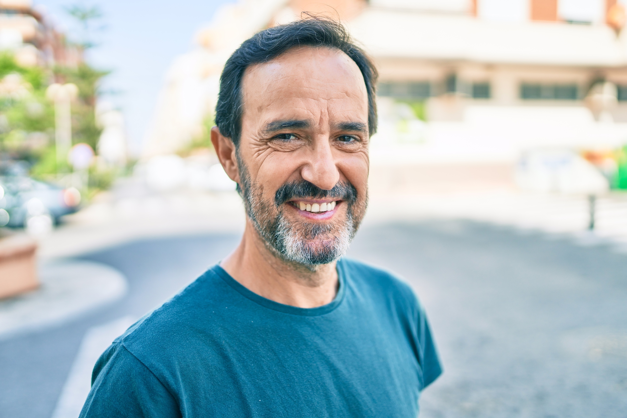 A middle-aged man with short dark hair and a graying beard is smiling while standing outdoors on a sunny day. He is wearing a simple blue T-shirt, and the background shows a blurred urban setting with buildings and cars.